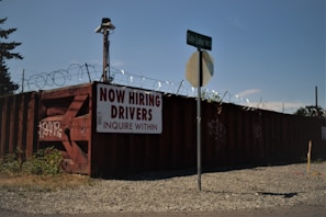 A barbed wire fence surrounds a perimeter with a large, rusty red shipping container that has graffiti on it. A sign on the container reads 'NOW HIRING DRIVERS INQUIRE WITHIN,' and a street sign for 'Tule Lake Rd S' is visible in front. The ground is covered with gravel and there is some overgrown grass.