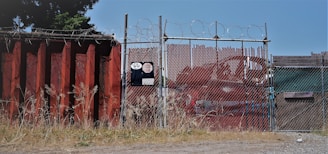 A chain-link fence topped with barbed wire stands secured around an industrial area. The fence is flanked by a large rusted metal wall on the left, with overgrown grass at its base. There is signage on the fence with visible numbers and logos. In the background, red metal containers can be seen.