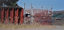 A chain-link fence topped with barbed wire stands secured around an industrial area. The fence is flanked by a large rusted metal wall on the left, with overgrown grass at its base. There is signage on the fence with visible numbers and logos. In the background, red metal containers can be seen.