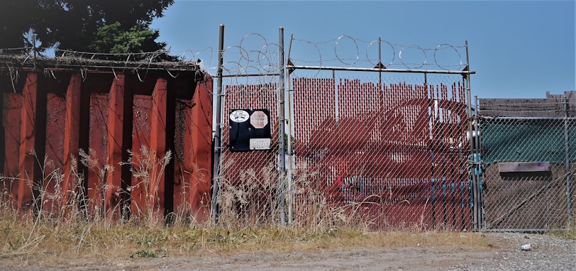 A chain-link fence topped with barbed wire stands secured around an industrial area. The fence is flanked by a large rusted metal wall on the left, with overgrown grass at its base. There is signage on the fence with visible numbers and logos. In the background, red metal containers can be seen.