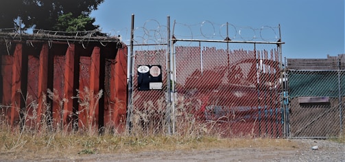 A chain-link fence topped with barbed wire stands secured around an industrial area. The fence is flanked by a large rusted metal wall on the left, with overgrown grass at its base. There is signage on the fence with visible numbers and logos. In the background, red metal containers can be seen.