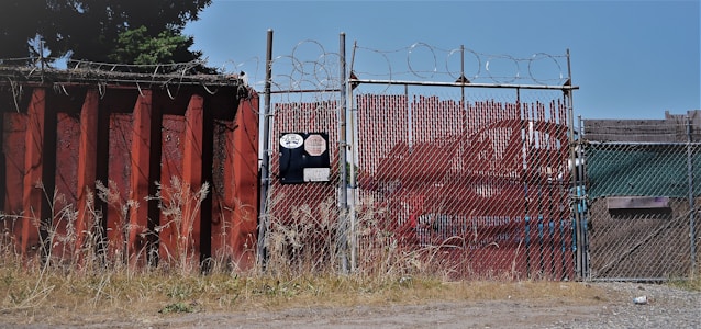 A chain-link fence topped with barbed wire stands secured around an industrial area. The fence is flanked by a large rusted metal wall on the left, with overgrown grass at its base. There is signage on the fence with visible numbers and logos. In the background, red metal containers can be seen.