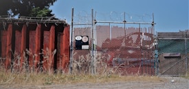 A chain-link fence topped with barbed wire stands secured around an industrial area. The fence is flanked by a large rusted metal wall on the left, with overgrown grass at its base. There is signage on the fence with visible numbers and logos. In the background, red metal containers can be seen.