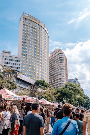 A lively street market is bustling with people walking and browsing stalls, which are covered with red and white striped canopies. Tall modern buildings with numerous windows loom in the background, while trees add greenery to the scene. The sky is partly cloudy, contributing to a pleasant and vibrant atmosphere.