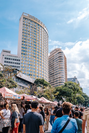 A lively street market is bustling with people walking and browsing stalls, which are covered with red and white striped canopies. Tall modern buildings with numerous windows loom in the background, while trees add greenery to the scene. The sky is partly cloudy, contributing to a pleasant and vibrant atmosphere.