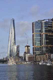 A modern waterfront cityscape featuring tall, reflective skyscrapers with a mix of architectural styles. The tallest building has a sleek, curved design, while others are rectangular with glass facades. Construction cranes are visible, suggesting ongoing development. Several boats and yachts are docked in the foreground, adding to the urban marina atmosphere.