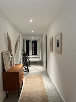 A narrow hallway with white walls and a polished concrete floor. Several ceiling lights illuminate the space. On the left, there is a wooden cabinet topped with decorative items including a vase with pampas grass. A bicycle is parked next to the cabinet. On the right wall, framed pictures are hung. A woven rug extends along the floor, leading towards a dark door at the end of the hallway.