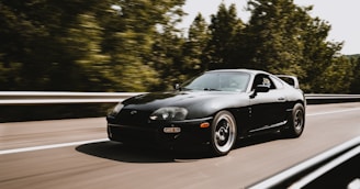 A sleek black sports car is driving at high speed on a highway. It appears to be on a smooth road flanked by lush green trees. The car's aerodynamic design and polished exterior suggest a sense of agility and power.