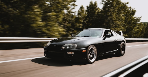 A sleek black sports car is driving at high speed on a highway. It appears to be on a smooth road flanked by lush green trees. The car's aerodynamic design and polished exterior suggest a sense of agility and power.