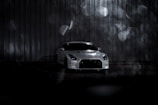 A white sports car is parked in front of a metallic corrugated backdrop, with dim lighting that casts soft shadows. The background is accentuated by out-of-focus circular light patterns creating a bokeh effect. The setting appears moody and atmospheric, with a focus on the sleek design of the car.