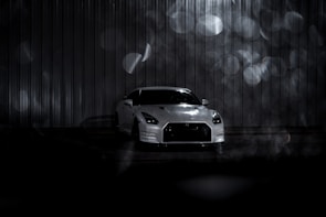 A white sports car is parked in front of a metallic corrugated backdrop, with dim lighting that casts soft shadows. The background is accentuated by out-of-focus circular light patterns creating a bokeh effect. The setting appears moody and atmospheric, with a focus on the sleek design of the car.