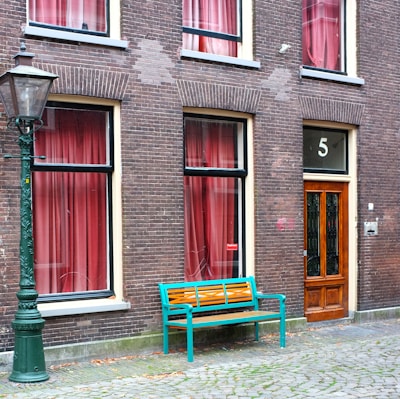 A brick building features multiple windows adorned with red curtains. A wooden door marked with the number 5 adds a distinct touch. A vibrant bench with a teal frame and wooden slats sits on the cobblestone path near an ornate street lamp.