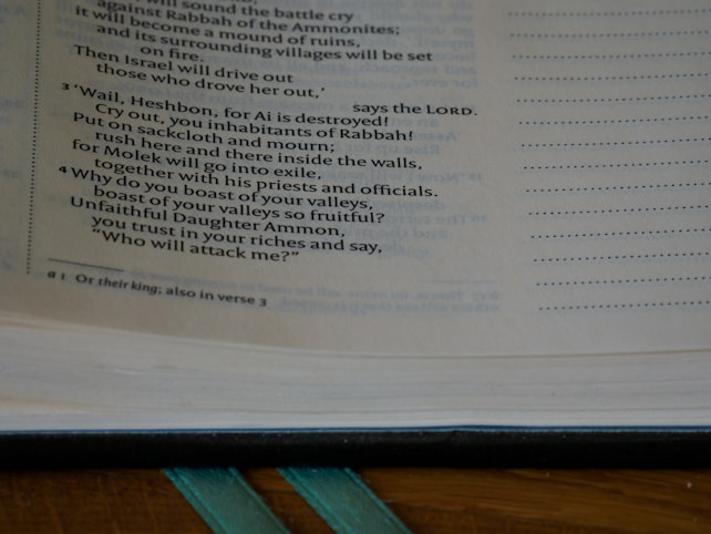 A close-up of an open book showing a page with biblical text and a footnote. The text is printed in black on an off-white page, with margins on both sides. A bookmark ribbon is visible at the bottom, and a portion of the book's cover edges can be seen.