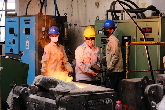 Three workers wearing safety helmets and protective gear stand in an industrial setting. Two of them observe while one holds a metal component near a machine involved with molten metal casting, emitting bright orange sparks. Control panels and heavy equipment are visible in the background.