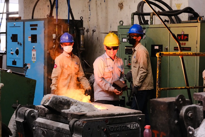 Three workers wearing safety helmets and protective gear stand in an industrial setting. Two of them observe while one holds a metal component near a machine involved with molten metal casting, emitting bright orange sparks. Control panels and heavy equipment are visible in the background.