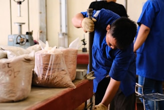 A worker dressed in a blue uniform is using a tool to press or mix contents in a bag. The workspace is filled with bags of material, possibly labeled with writing. The environment appears to be an industrial or laboratory setting with machinery in the background.