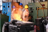 Three workers in a factory are wearing protective gear, such as helmets and goggles, while operating heavy machinery. Sparks are flying as one of them, wearing a yellow helmet, appears to be controlling a process involving heated metal.