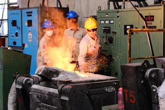 Three workers in a factory are wearing protective gear, such as helmets and goggles, while operating heavy machinery. Sparks are flying as one of them, wearing a yellow helmet, appears to be controlling a process involving heated metal.