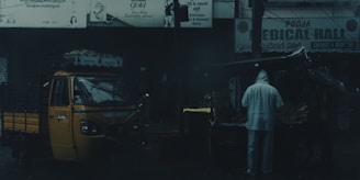A street scene in a dimly lit area featuring a yellow auto rickshaw parked next to a roadside stall. A person dressed in a raincoat is standing near the stall, which is likely to be selling goods. The background displays signage in multiple languages, and the atmosphere is moody and overcast.