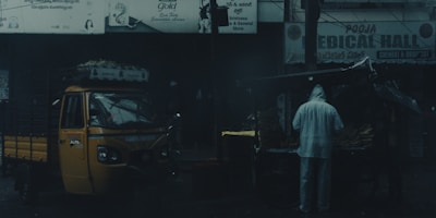 A street scene in a dimly lit area featuring a yellow auto rickshaw parked next to a roadside stall. A person dressed in a raincoat is standing near the stall, which is likely to be selling goods. The background displays signage in multiple languages, and the atmosphere is moody and overcast.