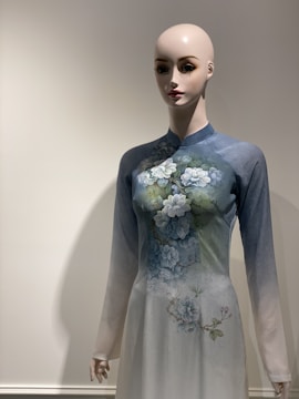 A mannequin is dressed in a traditional long-sleeved gown featuring a floral design with delicate blue and white flowers. The dress has a gradient that shifts from light blue at the shoulders to white towards the bottom. The mannequin is positioned against a plain, light-colored background that enhances the elegance of the attire.