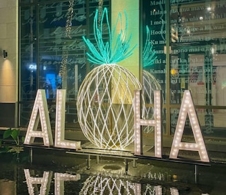 Large illuminated letters on a reflective surface spell out 'ALOHA' with a decorative pineapple shape in the background made from glowing lights. The scene is set in front of a glass wall with text in the background, creating a tropical and welcoming atmosphere.