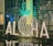 Large illuminated letters on a reflective surface spell out 'ALOHA' with a decorative pineapple shape in the background made from glowing lights. The scene is set in front of a glass wall with text in the background, creating a tropical and welcoming atmosphere.