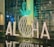 Large illuminated letters on a reflective surface spell out 'ALOHA' with a decorative pineapple shape in the background made from glowing lights. The scene is set in front of a glass wall with text in the background, creating a tropical and welcoming atmosphere.
