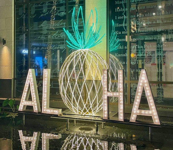 Large illuminated letters on a reflective surface spell out 'ALOHA' with a decorative pineapple shape in the background made from glowing lights. The scene is set in front of a glass wall with text in the background, creating a tropical and welcoming atmosphere.