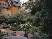 A serene garden surrounded by old buildings with rustic architecture. The garden is lush with greenery, colorful flowers, and well-maintained plants. There are several wooden chairs and a table on a paved area, suggesting a comfortable space for relaxation.