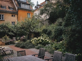 A serene garden surrounded by old buildings with rustic architecture. The garden is lush with greenery, colorful flowers, and well-maintained plants. There are several wooden chairs and a table on a paved area, suggesting a comfortable space for relaxation.