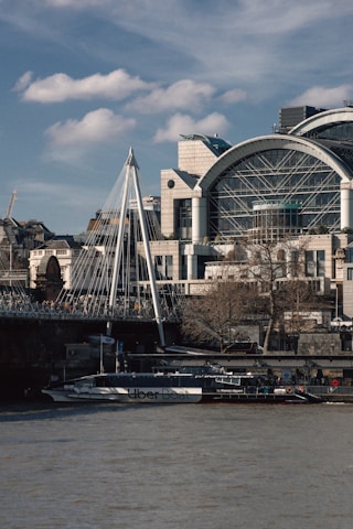 A modern architectural building with a large glass dome stands next to a suspension bridge. Below the bridge, there is an Uber Boat on the river, indicating a metropolitan setting with water transport.