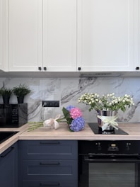 A modern kitchen counter with white cabinets above and blue drawers below. A bouquet of blue and pink hydrangeas lies on the wooden countertop, next to an elegant silver pot with white flowers. A black appliance is visible on the left, possibly a microwave, and two potted plants sit on top of it. The backsplash has a gray marble design.