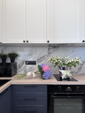A modern kitchen counter with white cabinets above and blue drawers below. A bouquet of blue and pink hydrangeas lies on the wooden countertop, next to an elegant silver pot with white flowers. A black appliance is visible on the left, possibly a microwave, and two potted plants sit on top of it. The backsplash has a gray marble design.
