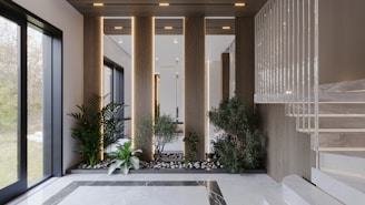 An elegant interior space featuring a modern staircase with marble steps. The area includes an arrangement of potted plants and small trees set among rocks, creating a serene garden-like atmosphere. Natural light streams in through large windows, highlighting the clean lines and sophisticated design.