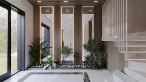 An elegant interior space featuring a modern staircase with marble steps. The area includes an arrangement of potted plants and small trees set among rocks, creating a serene garden-like atmosphere. Natural light streams in through large windows, highlighting the clean lines and sophisticated design.
