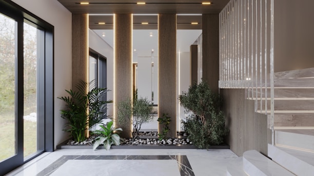 An elegant interior space featuring a modern staircase with marble steps. The area includes an arrangement of potted plants and small trees set among rocks, creating a serene garden-like atmosphere. Natural light streams in through large windows, highlighting the clean lines and sophisticated design.