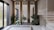 An elegant interior space featuring a modern staircase with marble steps. The area includes an arrangement of potted plants and small trees set among rocks, creating a serene garden-like atmosphere. Natural light streams in through large windows, highlighting the clean lines and sophisticated design.