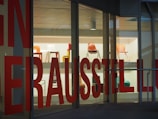Several colorful chairs are displayed on shelves inside a glass-walled building. Large red text is printed on the glass, partially obstructing the view of the chairs on display.