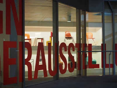 Several colorful chairs are displayed on shelves inside a glass-walled building. Large red text is printed on the glass, partially obstructing the view of the chairs on display.