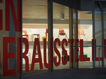 Several colorful chairs are displayed on shelves inside a glass-walled building. Large red text is printed on the glass, partially obstructing the view of the chairs on display.