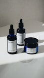 Three hemp-based skincare products are arranged on a smooth, white surface. Two are dropper bottles with dark blue glass and white labels, and one is a small jar with a screw-top lid, also featuring a white label. Natural lighting casts soft shadows on the surface.