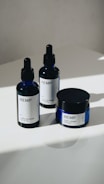 Three hemp-based skincare products are arranged on a smooth, white surface. Two are dropper bottles with dark blue glass and white labels, and one is a small jar with a screw-top lid, also featuring a white label. Natural lighting casts soft shadows on the surface.