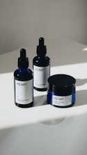 Three hemp-based skincare products are arranged on a smooth, white surface. Two are dropper bottles with dark blue glass and white labels, and one is a small jar with a screw-top lid, also featuring a white label. Natural lighting casts soft shadows on the surface.
