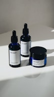 Three hemp-based skincare products are arranged on a smooth, white surface. Two are dropper bottles with dark blue glass and white labels, and one is a small jar with a screw-top lid, also featuring a white label. Natural lighting casts soft shadows on the surface.