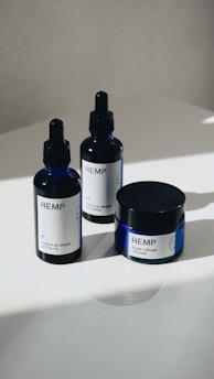 Three hemp-based skincare products are arranged on a smooth, white surface. Two are dropper bottles with dark blue glass and white labels, and one is a small jar with a screw-top lid, also featuring a white label. Natural lighting casts soft shadows on the surface.