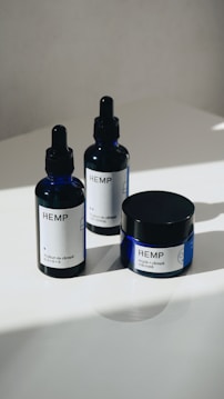 Three hemp-based skincare products are arranged on a smooth, white surface. Two are dropper bottles with dark blue glass and white labels, and one is a small jar with a screw-top lid, also featuring a white label. Natural lighting casts soft shadows on the surface.