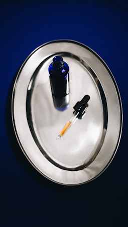 A blue glass bottle and a dropper with a yellow liquid are placed on a shiny, reflective silver tray. The background is a deep blue color, creating a contrast with the metallic surface.