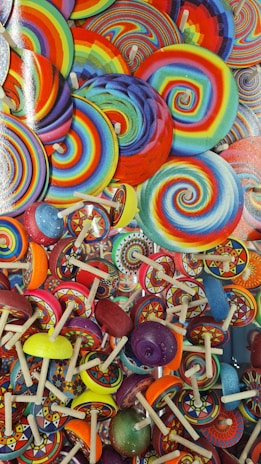 A vibrant collection of colorful spinning tops and lollipops featuring swirling patterns and a wide array of colors including red, blue, yellow, and green. The designs are intricate, with concentric circles and spiral motifs that give a playful and lively appearance to the scene.