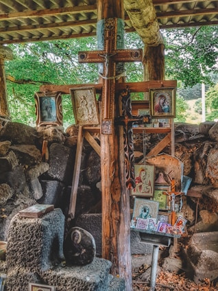 A collection of religious icons, paintings, and a large wooden cross are arranged against a rustic backdrop of wooden beams and stone. The setting appears to be outdoors or in a partially enclosed space, with surrounding greenery visible. The various images depict Christian religious figures and scenes, carefully displayed and propped up.